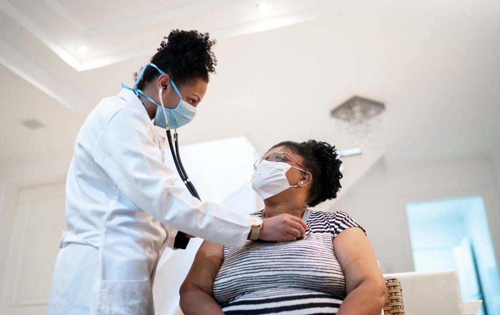 doctor listening to patient's heartbeat during home visit wearing face mask